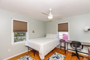 A bedroom with a bed, desk, chair, ceiling fan, and windows with blinds.