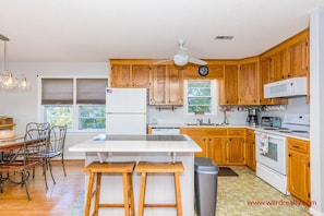 A kitchen with wooden cabinets, a white refrigerator, a microwave, a stove, and a ceiling fan. There is a window with a view of trees and a dining area with a table and chairs.