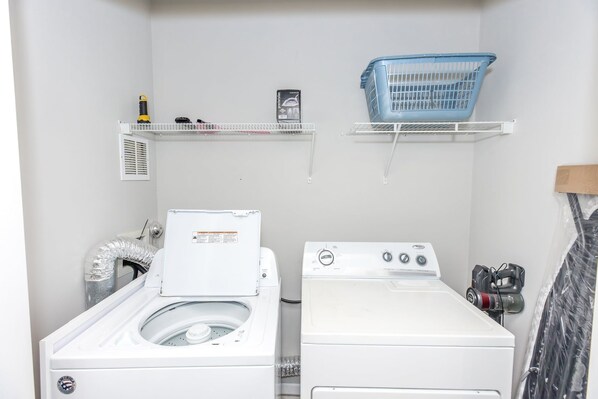 A laundry room with two washing machines, a blue laundry basket, and a shelf with cleaning supplies.