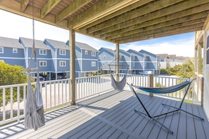 A covered outdoor deck with a hammock, wooden railings, and a view of blue residential buildings. - Shore Is Nice - 4BR Townhouse with Easy Beach Access! Pet Friendly! (Surf City)