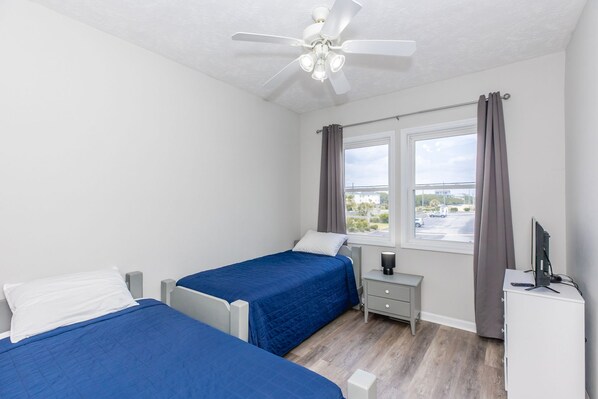 A bedroom with two beds, a ceiling fan, a window with curtains, a bedside table, and a TV stand.