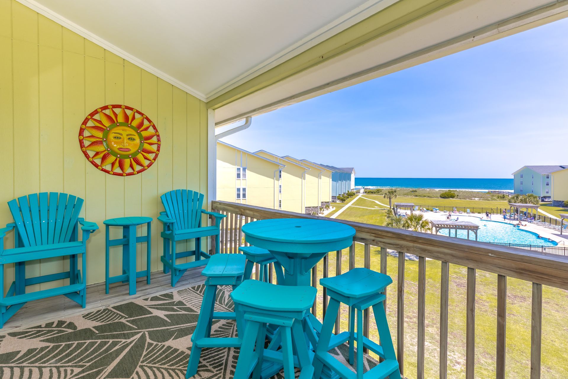 Un balcone con sedie e tavoli color turchese, un decoro solare e vista sulla spiaggia e sulla zona della piscina.