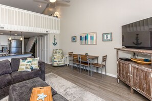 Ein Wohnzimmer mit hohen Decken, einem Essbereich mit Holztisch und -stühlen, einem grauen Sofa mit Zierteilen und einem Fernseher auf einer Holzkommode.