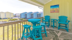 A balcony with turquoise chairs and stools, a patterned rug, and a painting of a bicycle on the wall. - The Sunny Surf - Top Floor Surf Condo - 2 BRs, 2 BA! (Surf City)