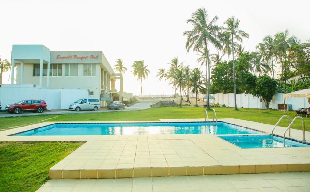 Una piscina con plataforma de azulejos, palmeras y un edificio que dice "Emerald Beach Resort".