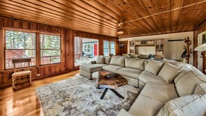 A spacious living room with a large sectional sofa, wooden ceiling, and large windows offering a view of the outdoors.