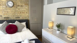 A modern hotel room with a brick wall, a bed with white bedding and a red pillow, a bedside table with two lamps, and a framed artwork on the wall. - Modern en-suite Studio in the Heart of Mill Road (Cambridge)