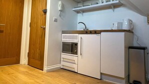 A compact kitchen with a microwave, oven, and sink, a wooden door, and a shelf with dishes. - Modern ensuite Studio Retreat off Vibrant Mill Rd (Cambridge)