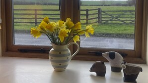 Une fenêtre donnant sur un paysage rural, un vase rempli de fleurs jaunes et deux statuettes d’animaux en céramique posées sur un rebord de fenêtre.