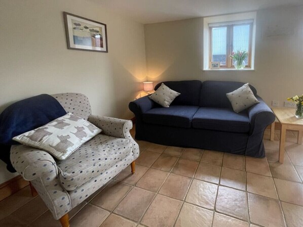 A living room with a patterned armchair, a dark blue sofa, a small table with a lamp, and a window with a view.