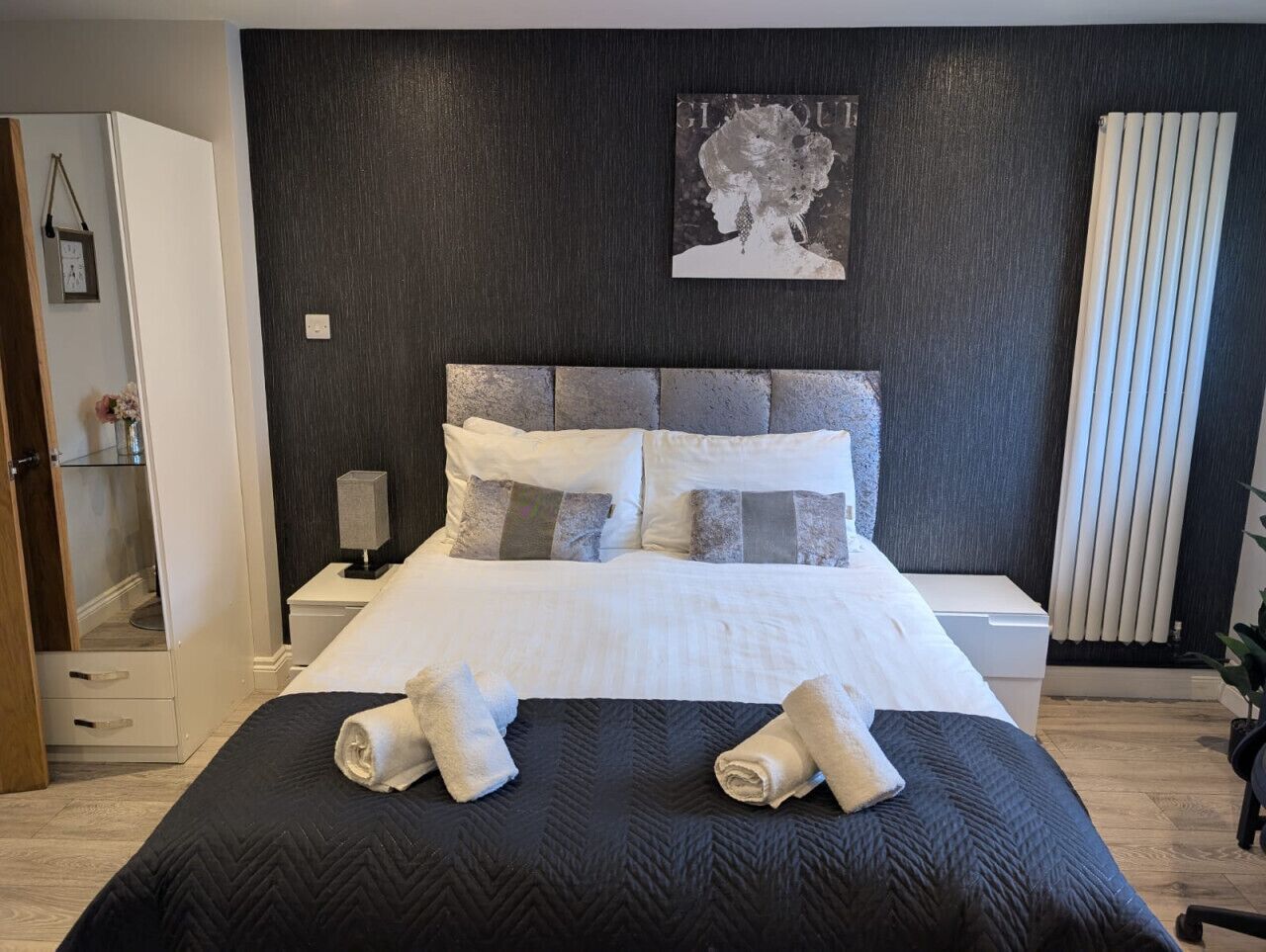 A neatly made bed with rolled towels, a bedside table with a lamp, a dark wall with a framed picture, and a radiator.
