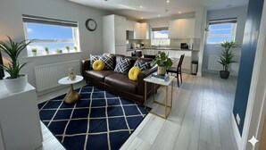 A modern apartment with a living room, kitchen, and dining area. It features a brown leather sofa, a blue and white rug, and potted plants.