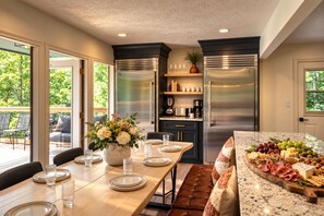 A modern kitchen with stainless steel appliances, a dining area with a wooden table set for four, and a view of the outdoors through large windows.