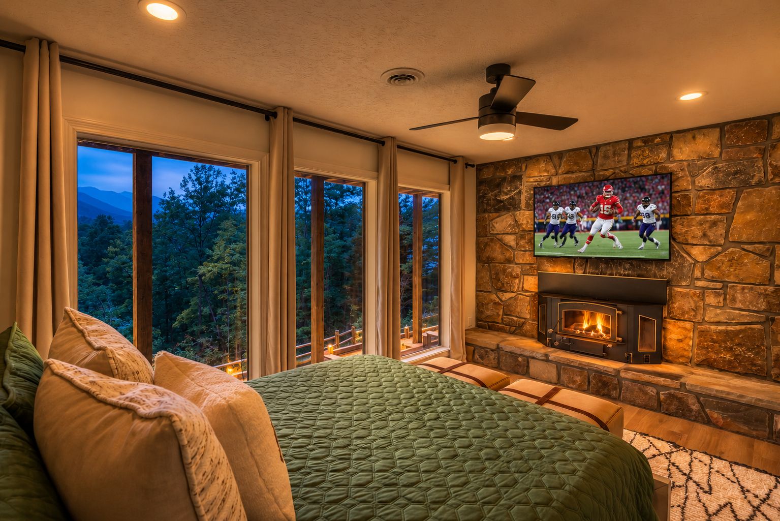A bedroom with a large bed, a stone fireplace, a television mounted on the wall, and large windows with a view of trees.