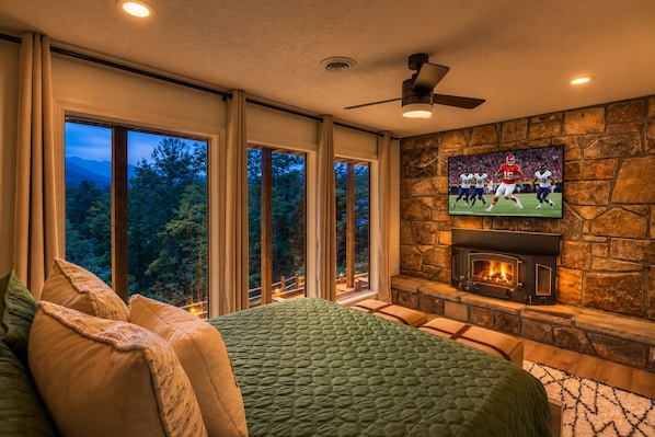 A bedroom with a large bed, a stone fireplace, a television mounted on the wall, and large windows with a view of trees.