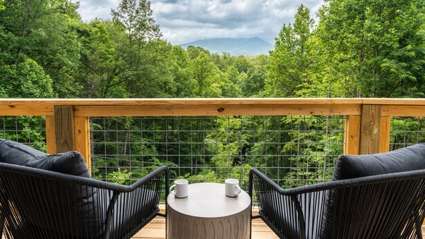 Una terraza de madera con dos sillas de mimbre negro y una mesa redonda, con vistas a un bosque frondoso y una cordillera de montañas.