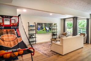 A living room with a basketball hoop, a large flat-screen TV, a sofa, and a window with curtains. - Stunning Luxury Retreat With Sweeping Mountain Views!  Something for Everyone! (Gatlinburg)