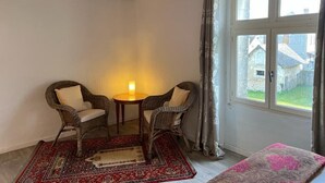 Ein Zimmer mit zwei Korbstühlen, einem kleinen Tisch, einem roten Teppich mit Muster und Blick durch ein Fenster nach draußen.