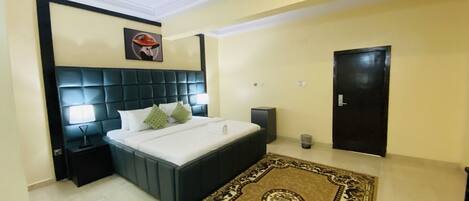 A hotel room with a large bed, a headboard, a bedside table, a lamp, a trash bin, a door, and a patterned rug.