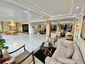 Um hall amplo com piso de mármore, área de estar central com sofá e poltronas, e um candelabro.