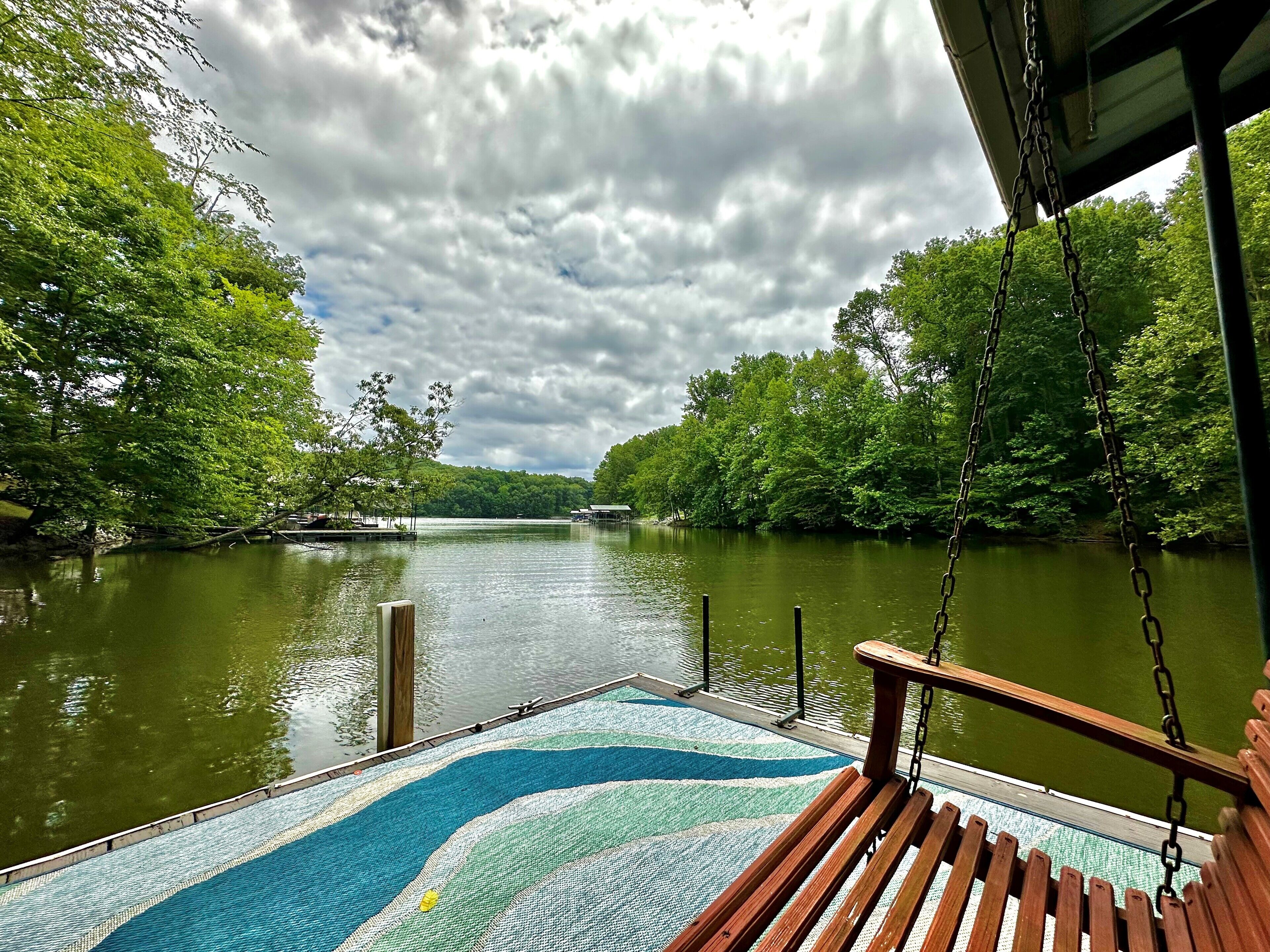 Ein Bootsdeck mit einer hölzernen Bank und einem gemusterten Teppich, mit Blick auf einen ruhigen Fluss, der von üppigem Grün umgeben ist.