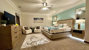 Een slaapkamer met een bed, twee fauteuils, een dressoir, een plafondventilator, een televisie en een schilderij aan de muur.