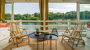 Una habitación con vistas a un lago, dos sillones de mimbre, una mesita redonda y un sofá blanco.