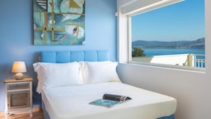 A bedroom with a bed, a nightstand, a lamp, a painting, and a window with a view of the sea and hills. - Villa Ventalia with magnificent panoramic sea view! (Lefkada)