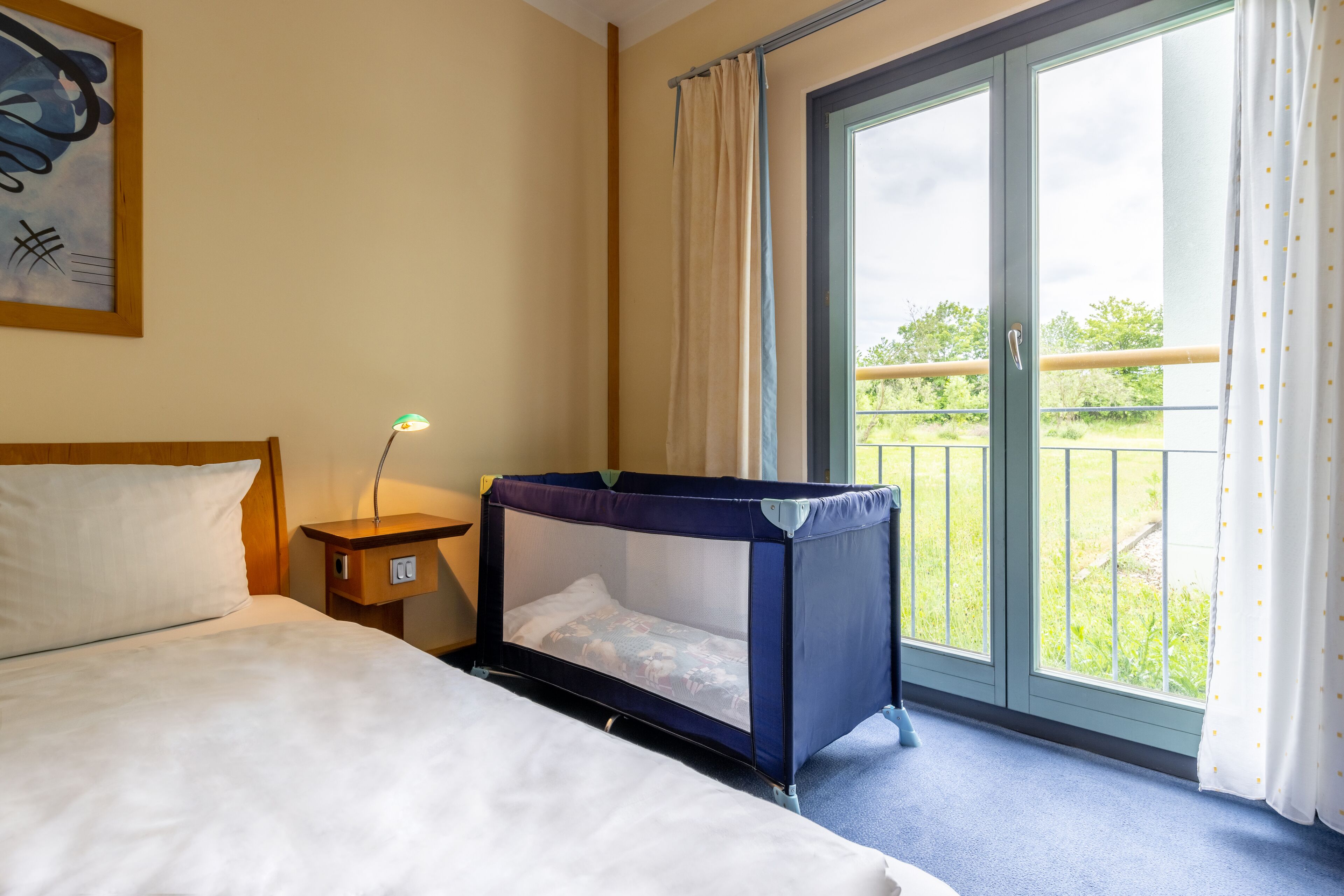 Een hotelkamer met een bed, een klein nachtkastje met een lamp, een raam met gordijnen en een balkon met uitzicht op groen.