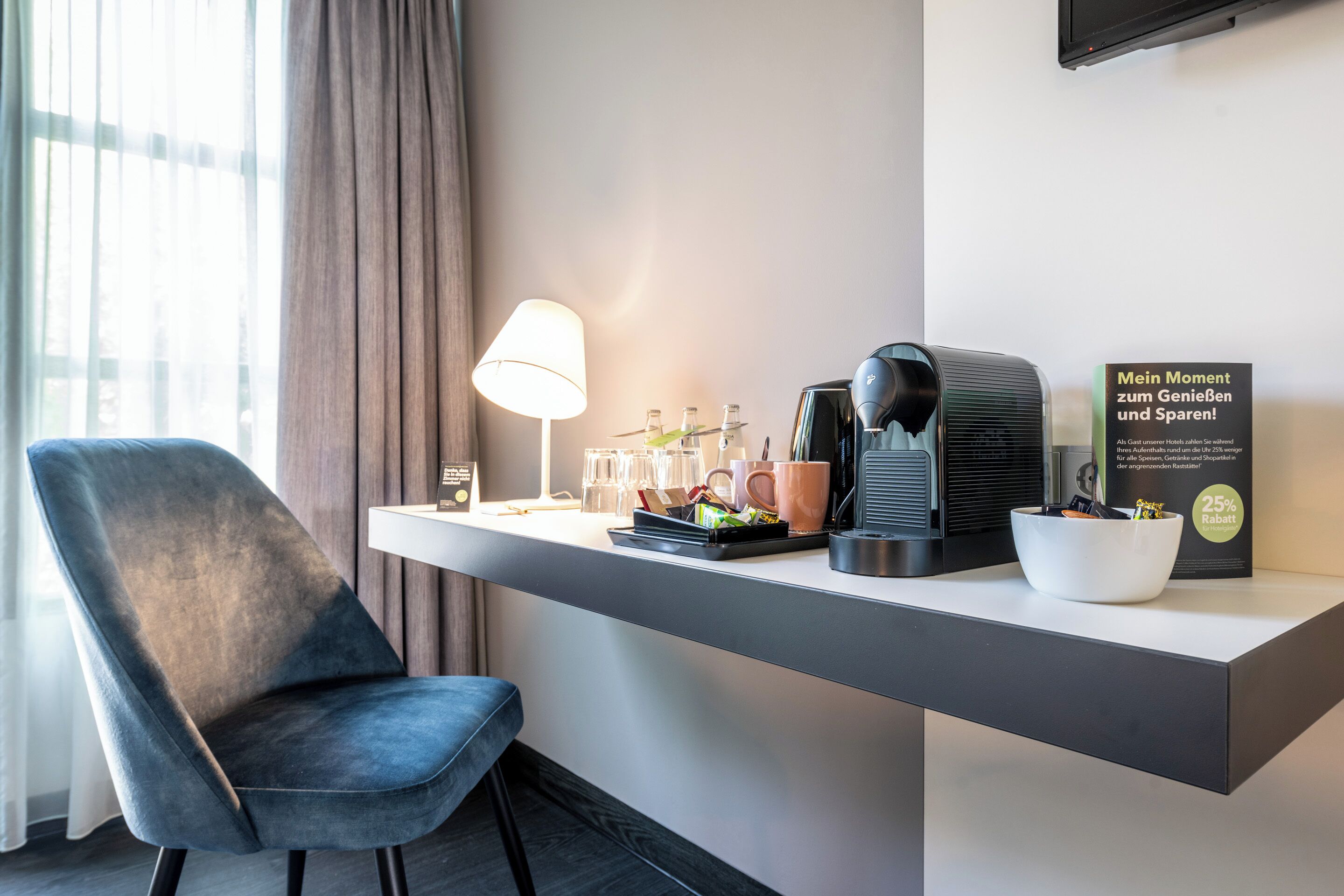 Een moderne hotelkamer met een donkerblauwe stoel, een wit bureau, een lamp, een koffiezetapparaat, een schaal en een bord waarop een korting van 25% wordt aangeboden.