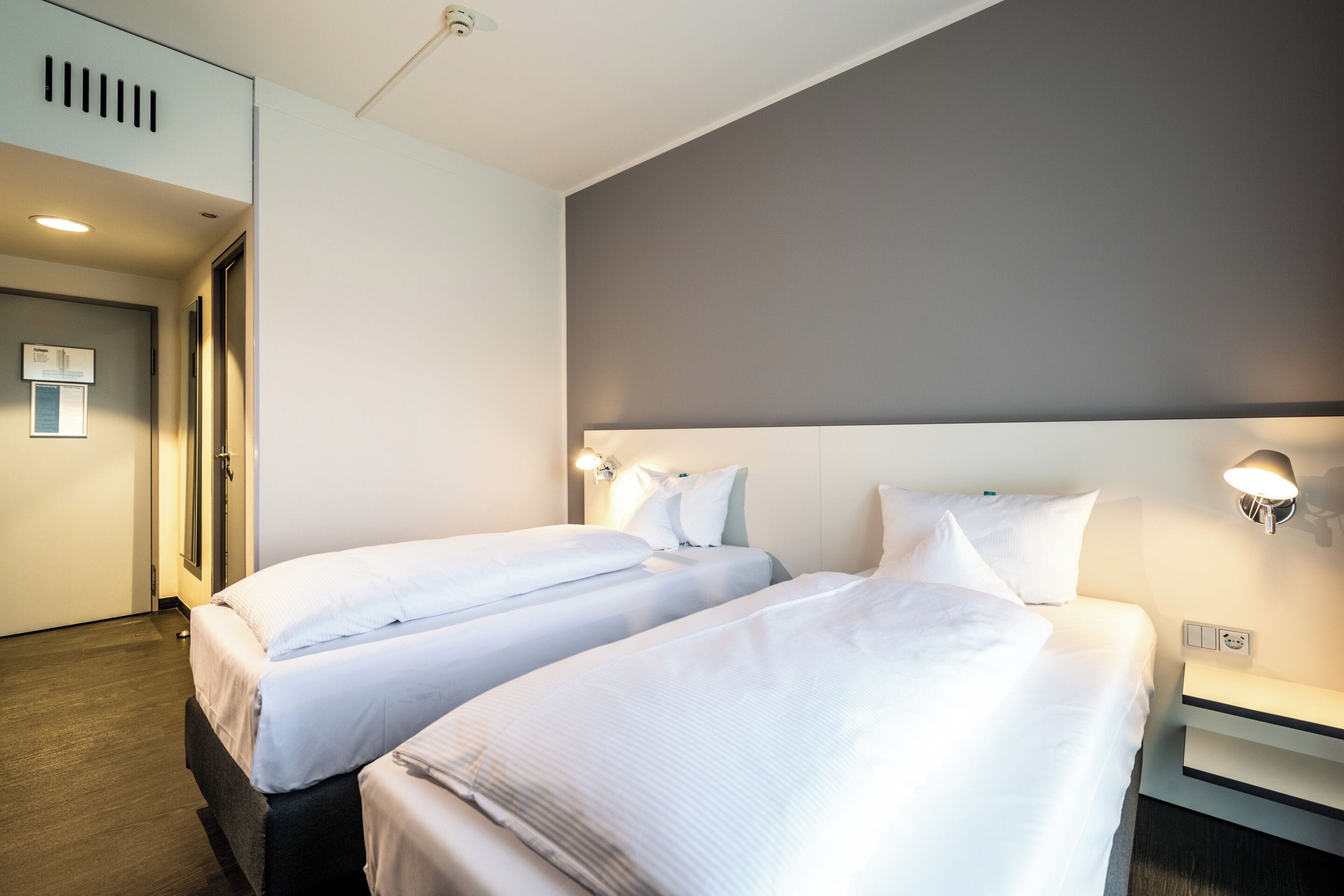 A hotel room with two beds, a bedside table, a nightstand, a wall-mounted light, and a door.