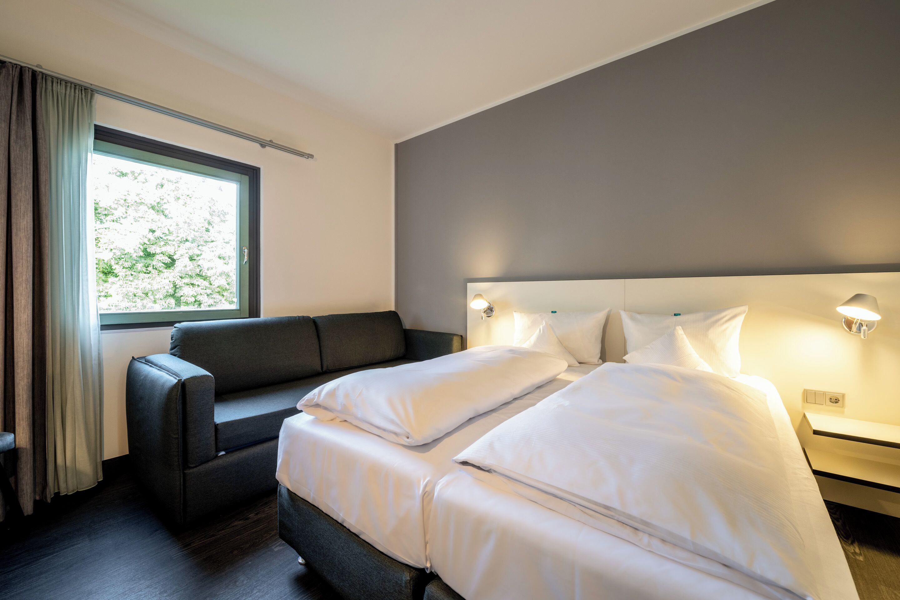 A modern hotel room with a large bed, a sofa, a window with curtains, and bedside lamps.