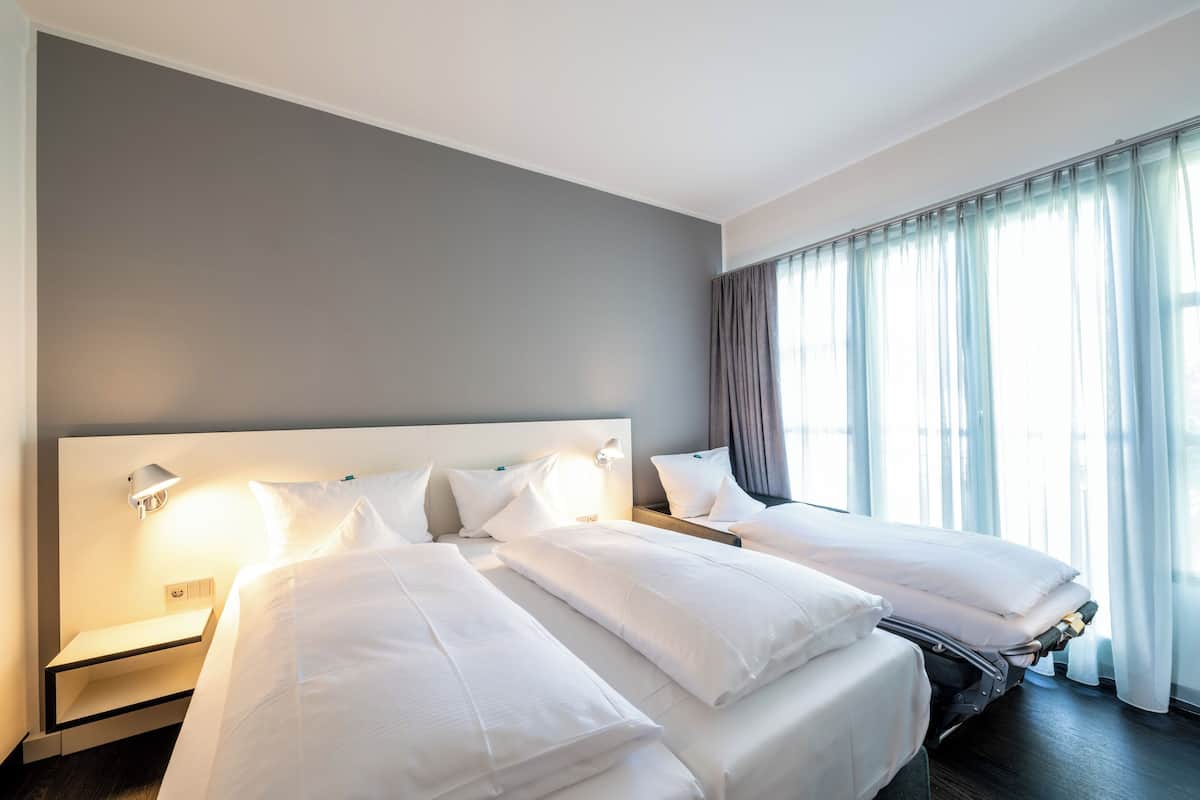 Ein Hotelzimmer mit zwei Betten, einem Nachttisch, einer Lampe und einem großen Fenster mit Vorhängen.