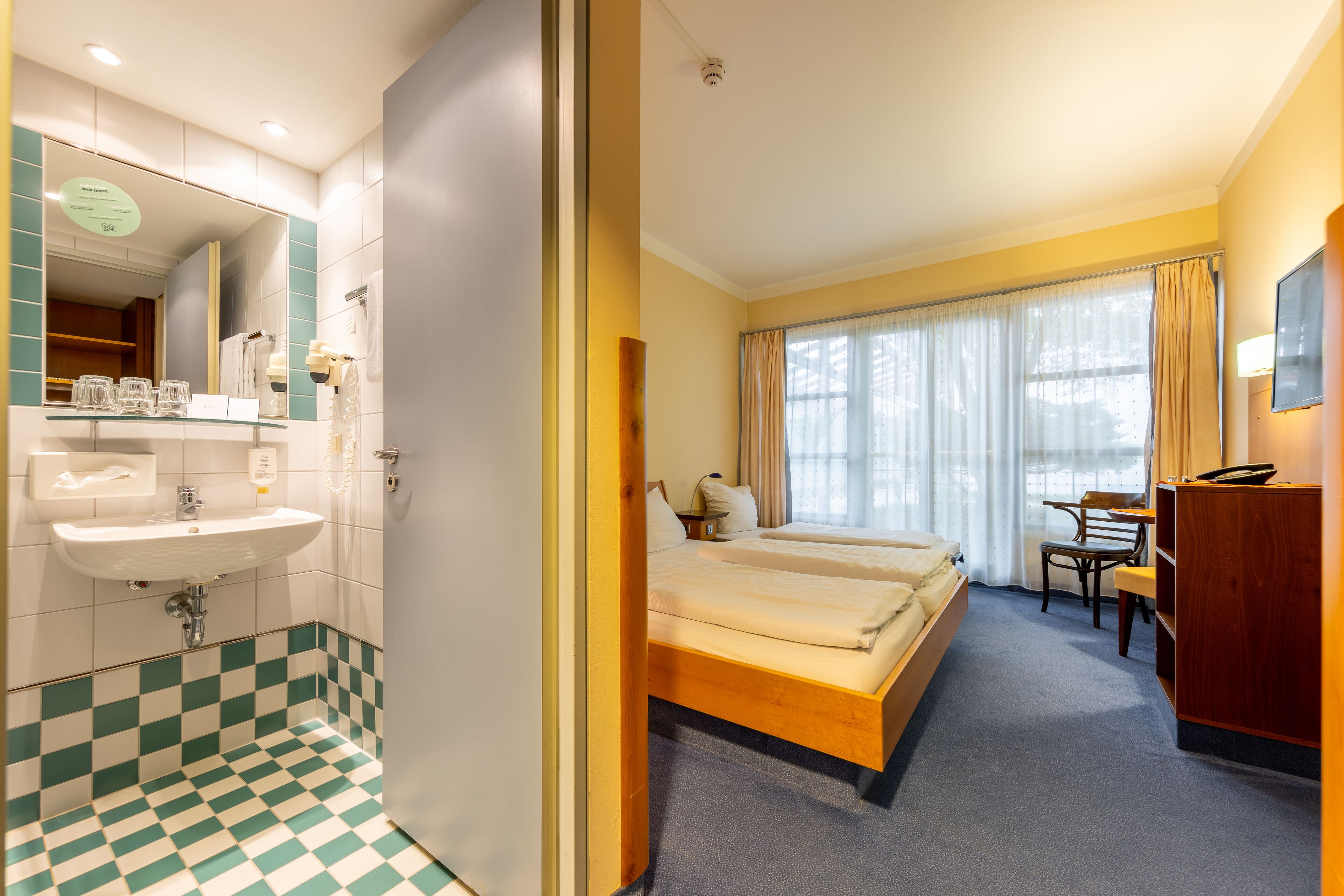 A hotel room with a checkered floor, a bed, a desk, a chair, a TV, and a bathroom with a sink and mirror.