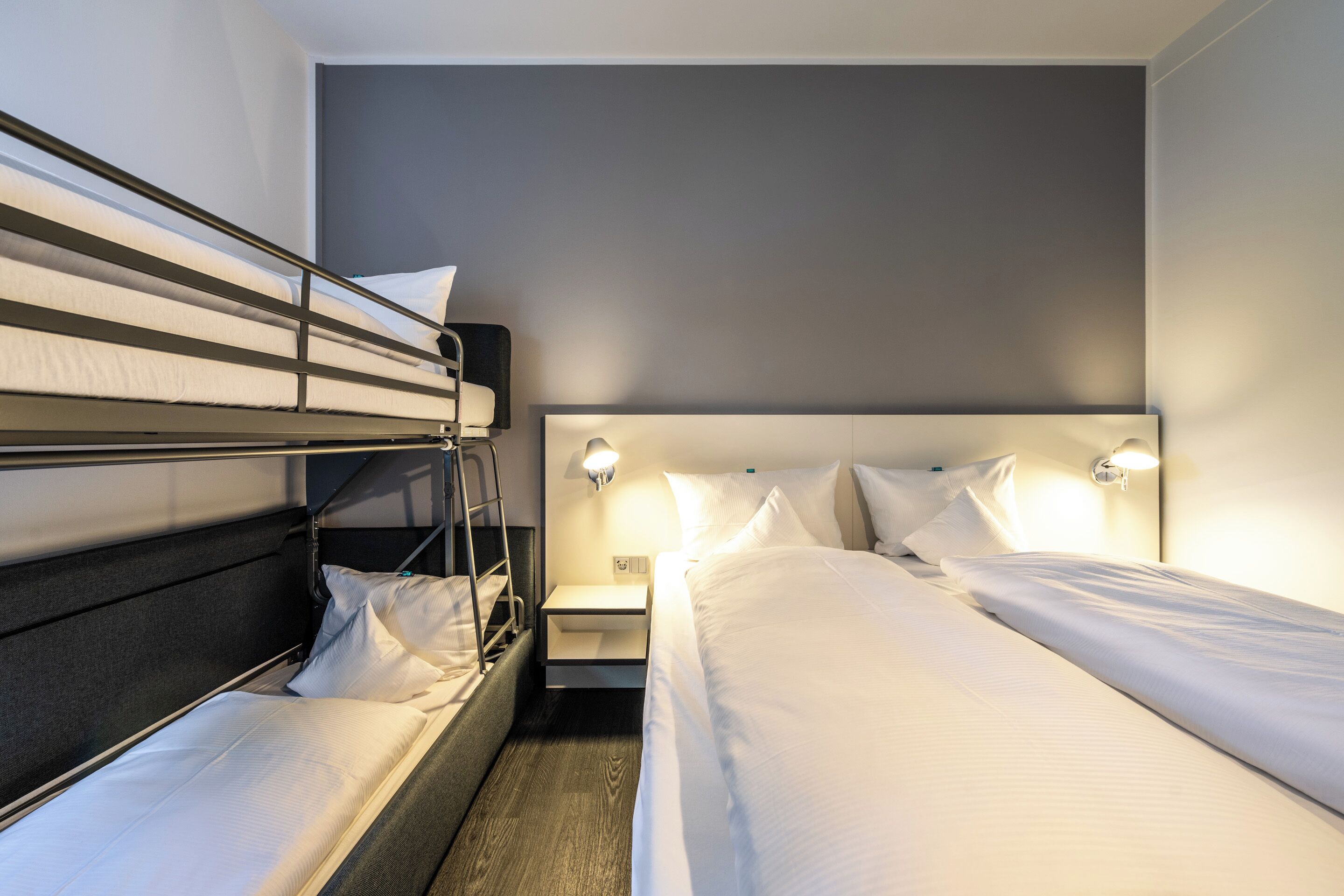 A hotel room with a bunk bed, a single bed, a nightstand with a lamp, and a wall-mounted light fixture.