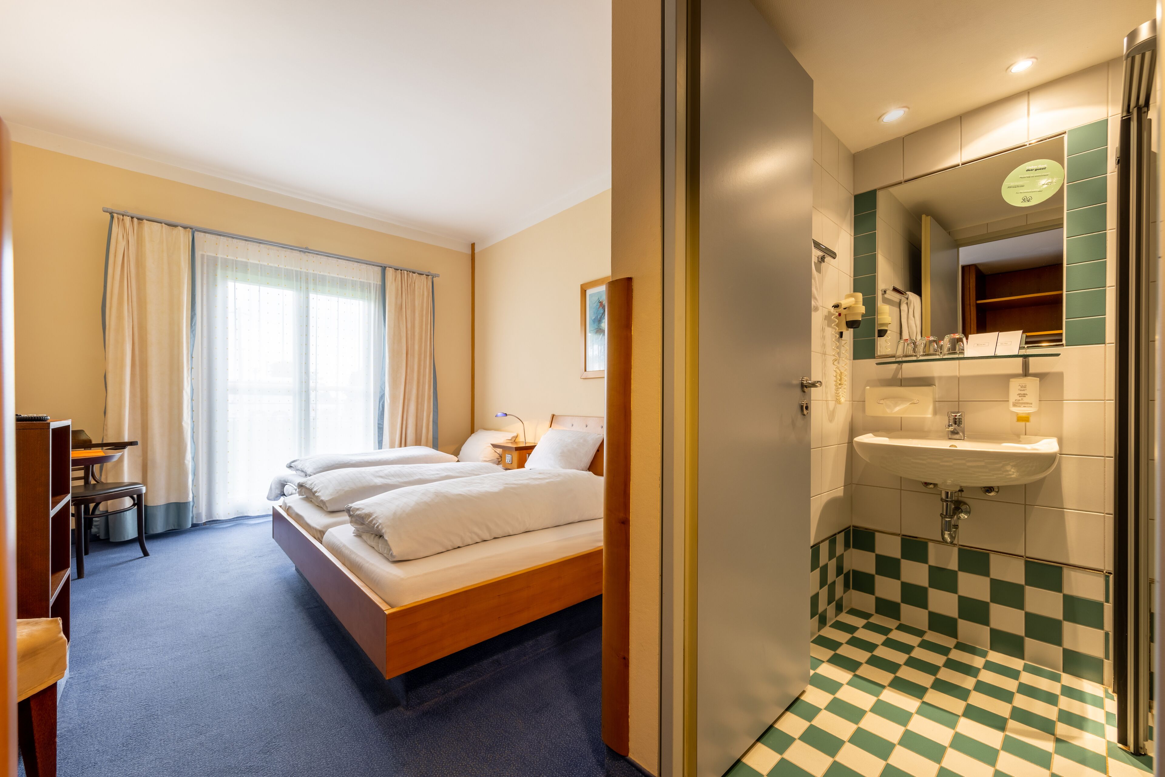 Habitación de hotel con cama, escritorio, silla, baño con lavabo y espejo, y baldosas de suelo a cuadros.