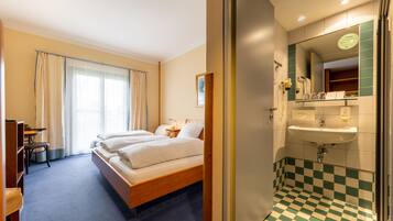 Habitación de hotel con cama, escritorio, silla, baño con lavabo y espejo, y baldosas de suelo a cuadros.