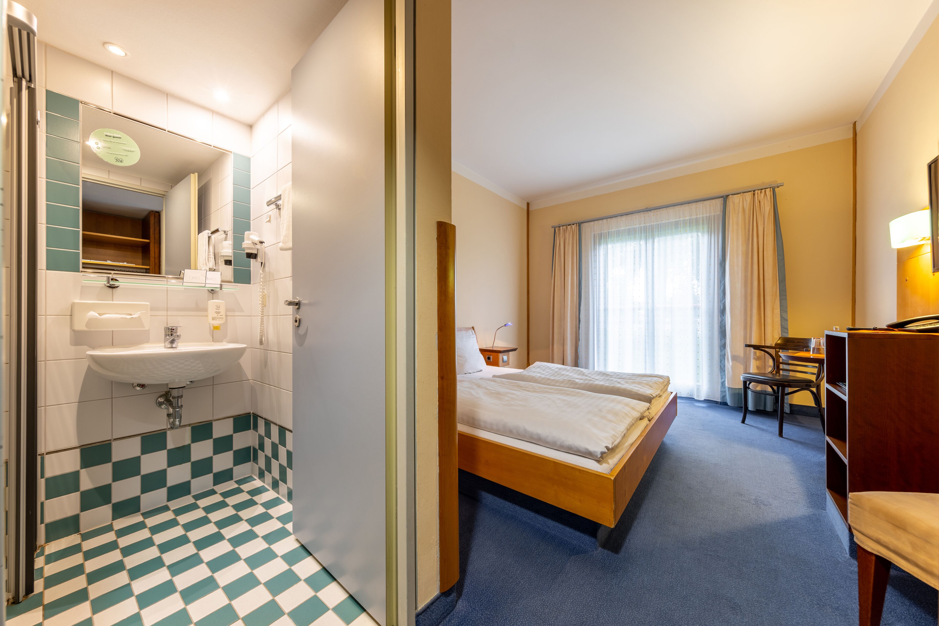 Ein Hotelzimmer mit Schachbrettboden, einem Bett, einem Schreibtisch, einem Sessel, einem Spiegel und einem Waschbecken.