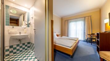 Ein Hotelzimmer mit Schachbrettboden, einem Bett, einem Schreibtisch, einem Sessel, einem Spiegel und einem Waschbecken.