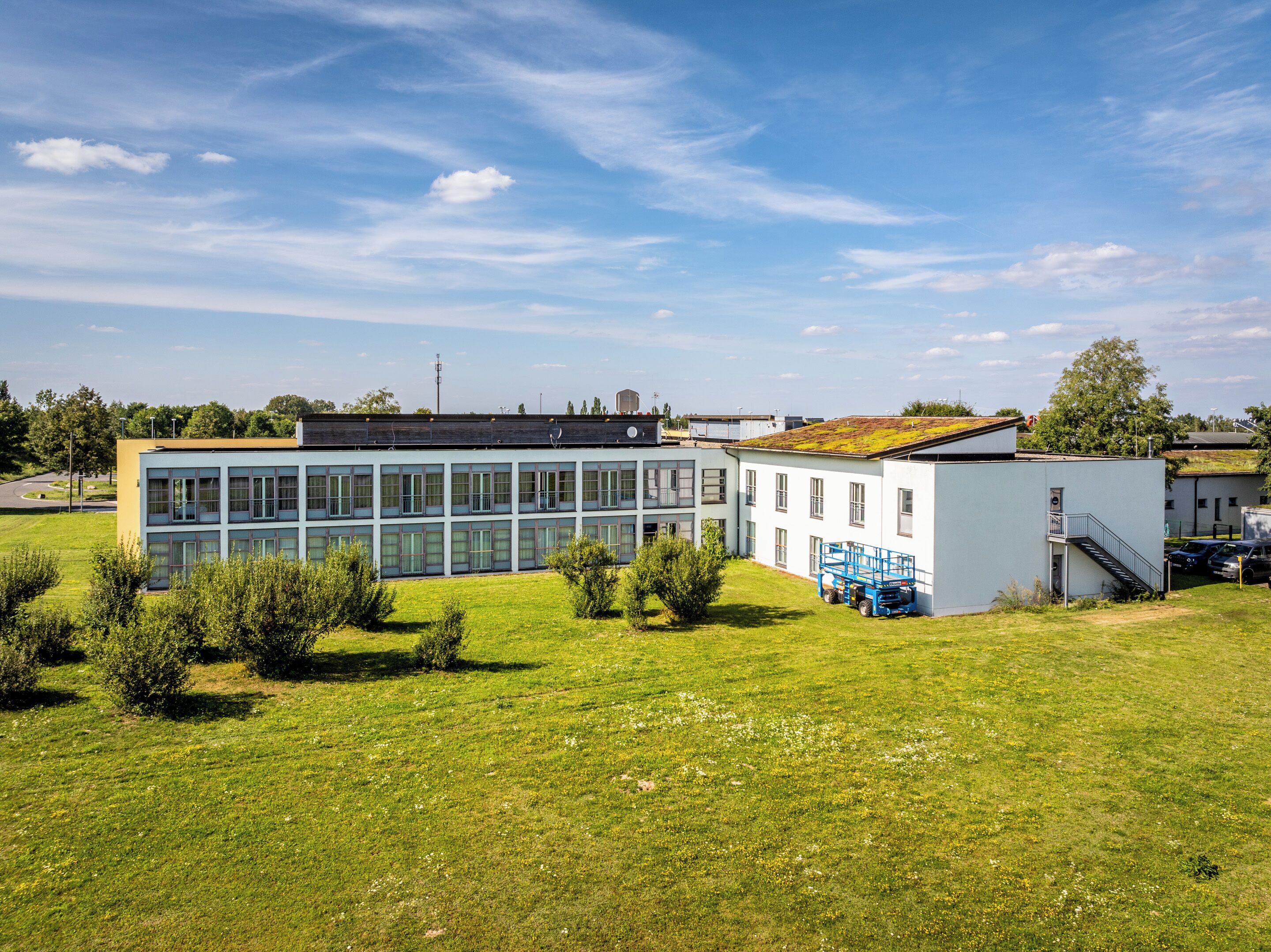Un bâtiment moderne doté de grandes fenêtres et d’un toit plat, entouré d’un champ verdoyant et de quelques arbres.