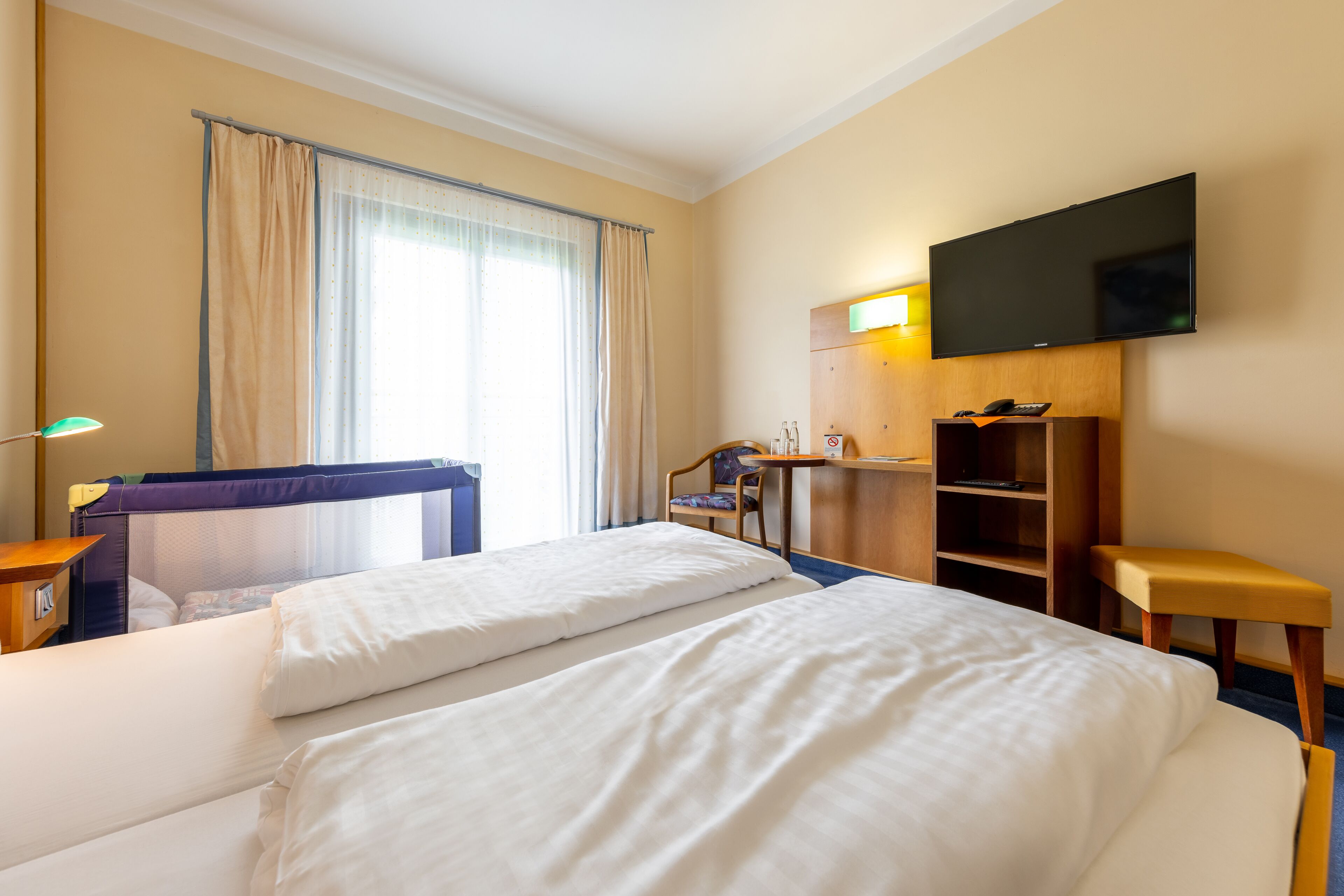 Hotellihuone, jossa on kaksi sänkyä, työpöytä, tuoli, televisio ja ikkuna, jossa on verhot.