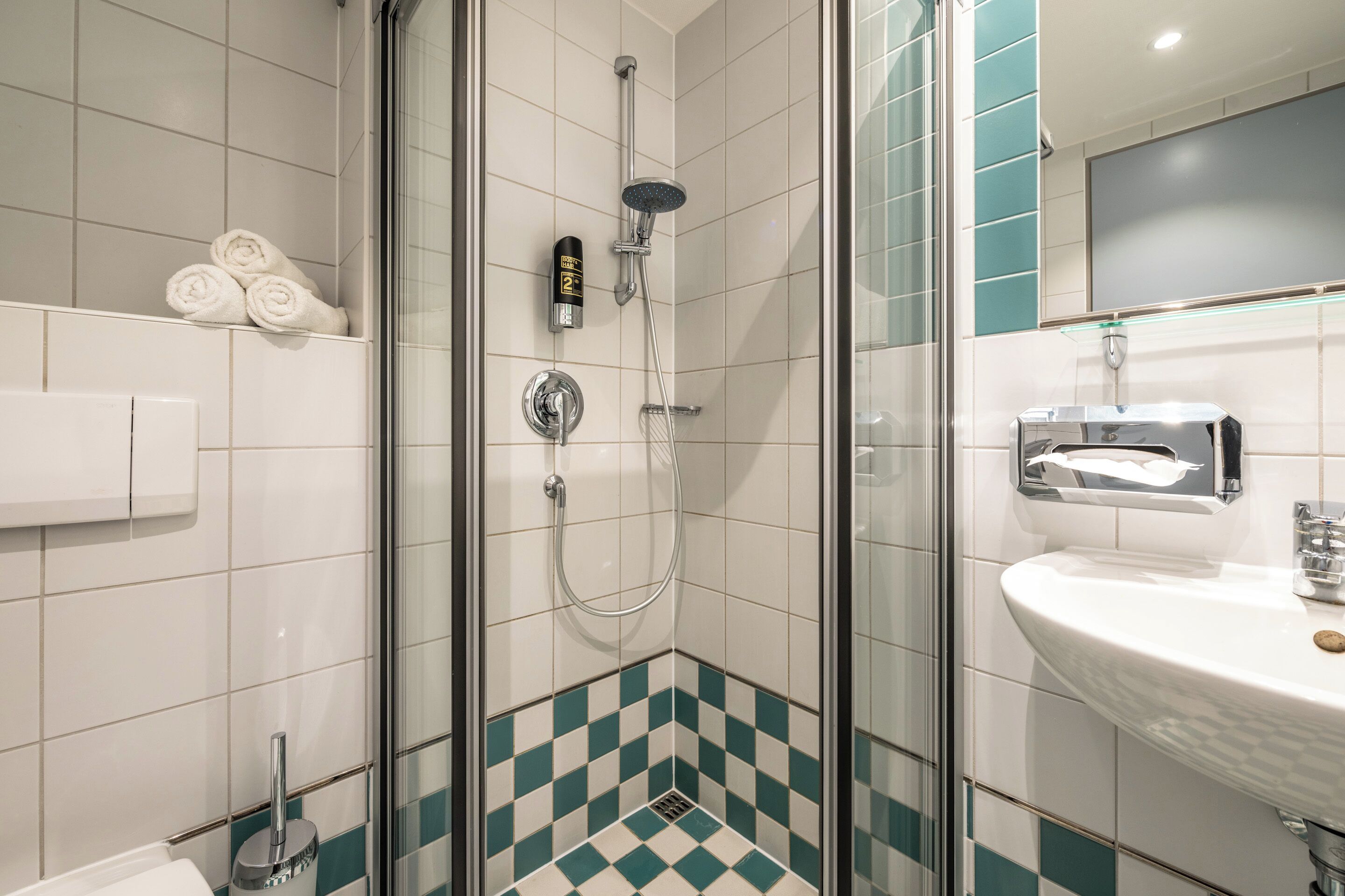 Une salle de bain moderne avec une douche à parois de verre, un lavabo blanc doté d’un distributeur de savon et un porte-serviettes avec des serviettes soigneusement pliées.