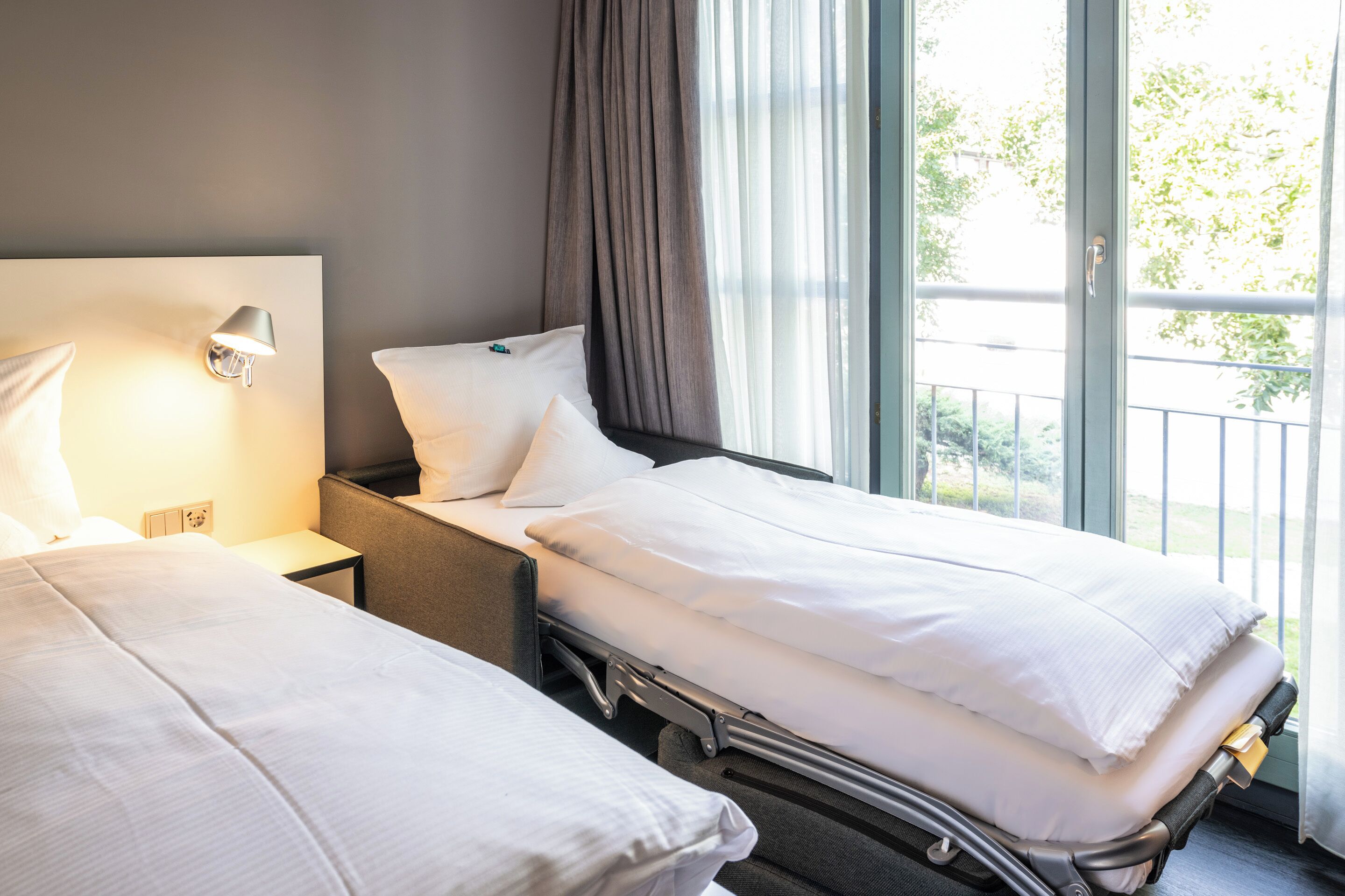 A hotel room with two beds, a bedside table with a lamp, and a view of greenery through the window.