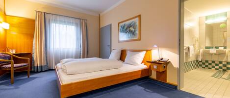 A hotel room with a large bed, a desk with a chair, a bedside table with a lamp, and a bathroom visible through an open door.