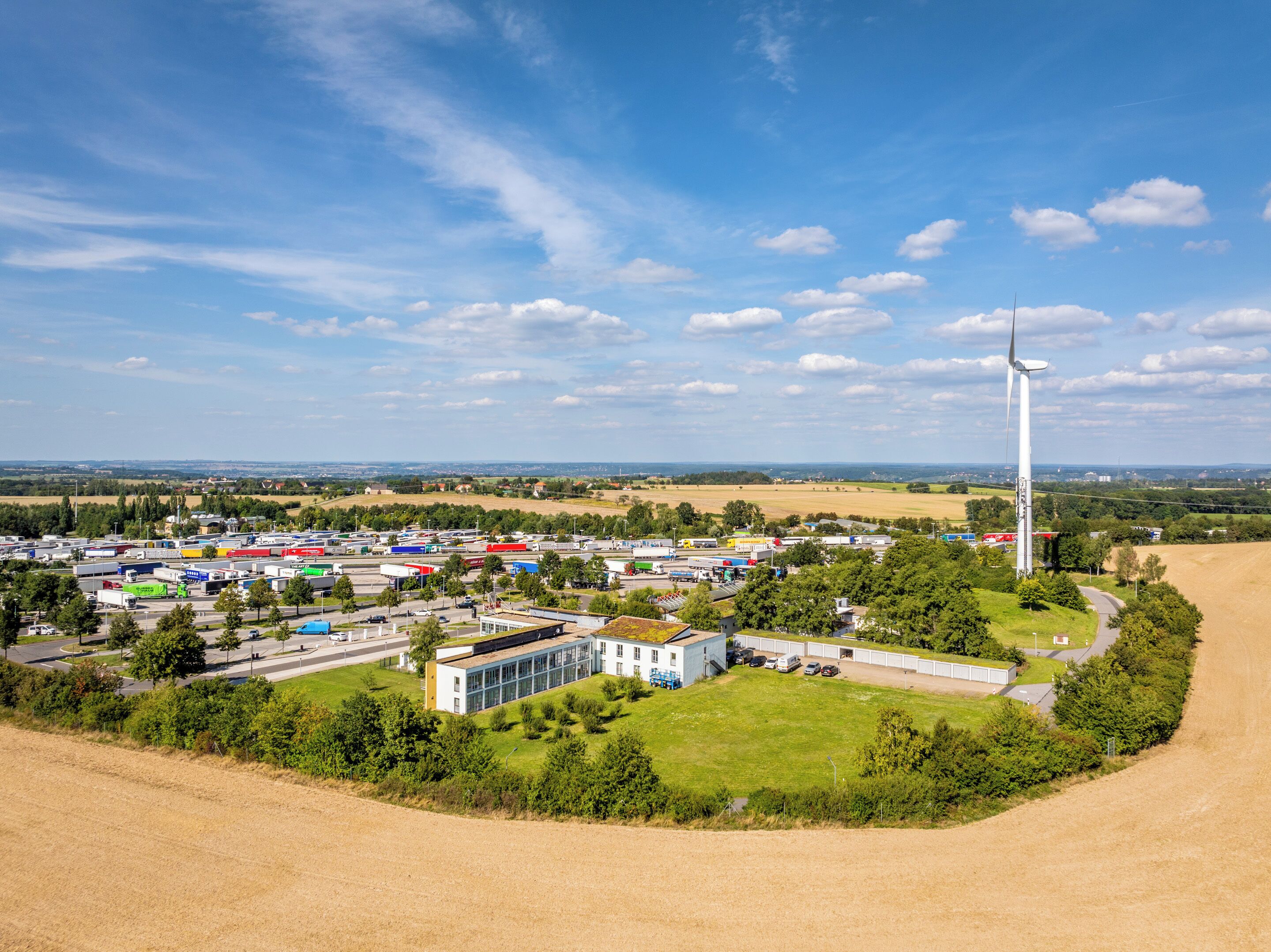 Ein weitläufiges Industriegebiet mit mehreren Gebäuden, einer Windkraftanlage und angrenzenden Feldern.