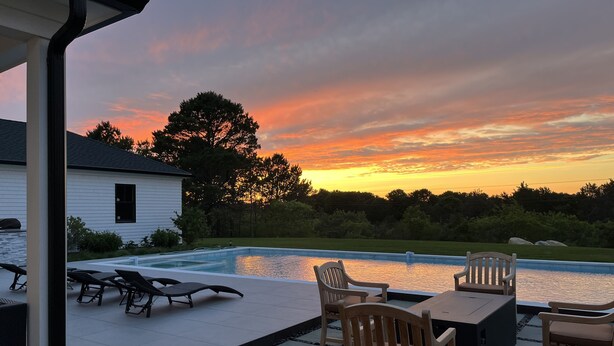 Piscina con sillas de descanso y una mesa, rodeada por un patio de piedra y vistas a un atardecer.