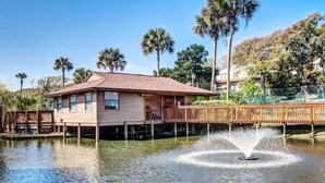 Una casa con un muelle que se adentra en un cuerpo de agua, rodeada de palmeras y un cielo despejado.