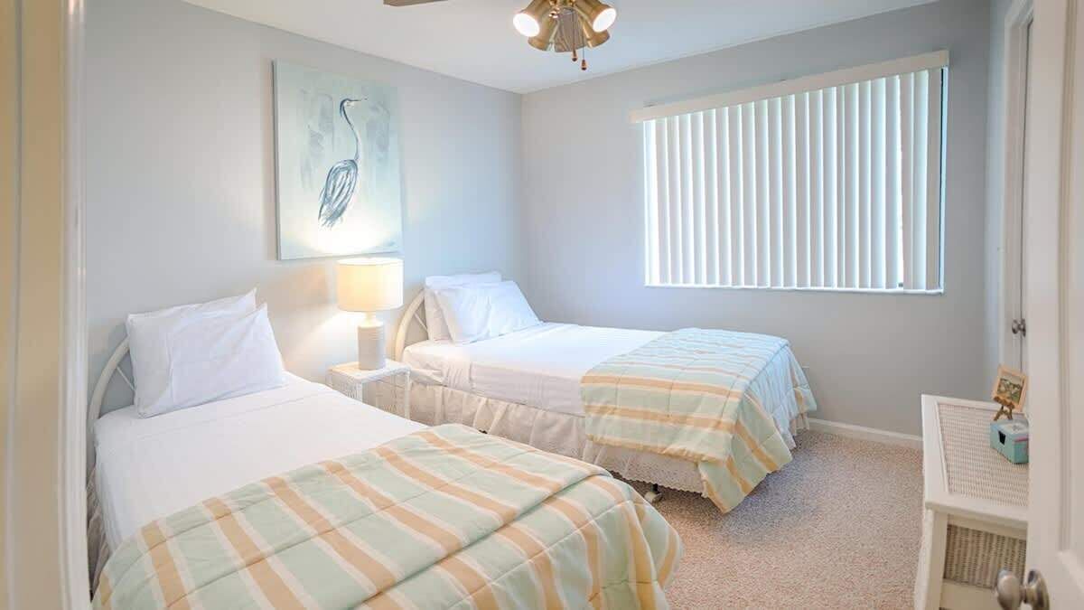 A bedroom with two beds, a window with blinds, a ceiling fan, and a painting on the wall.