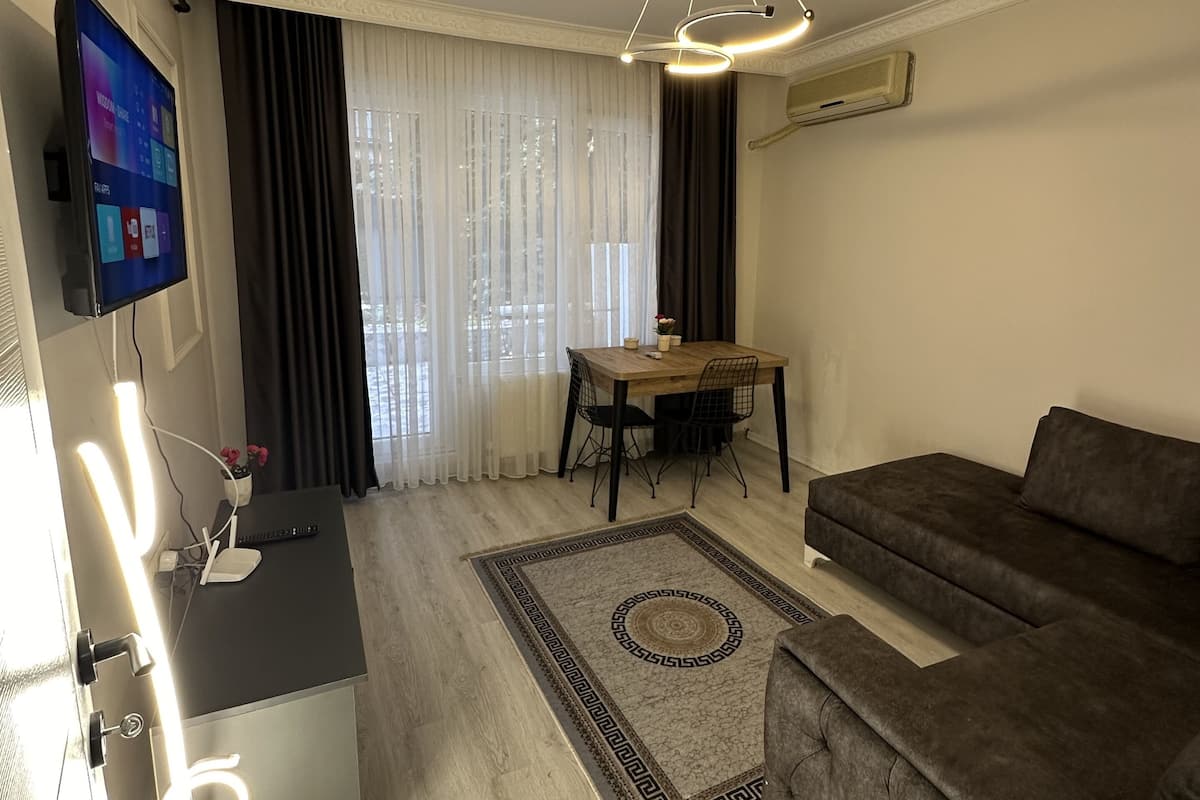 A modern living room with a flat-screen TV, a dining table with chairs, a brown sofa, and a patterned rug.
