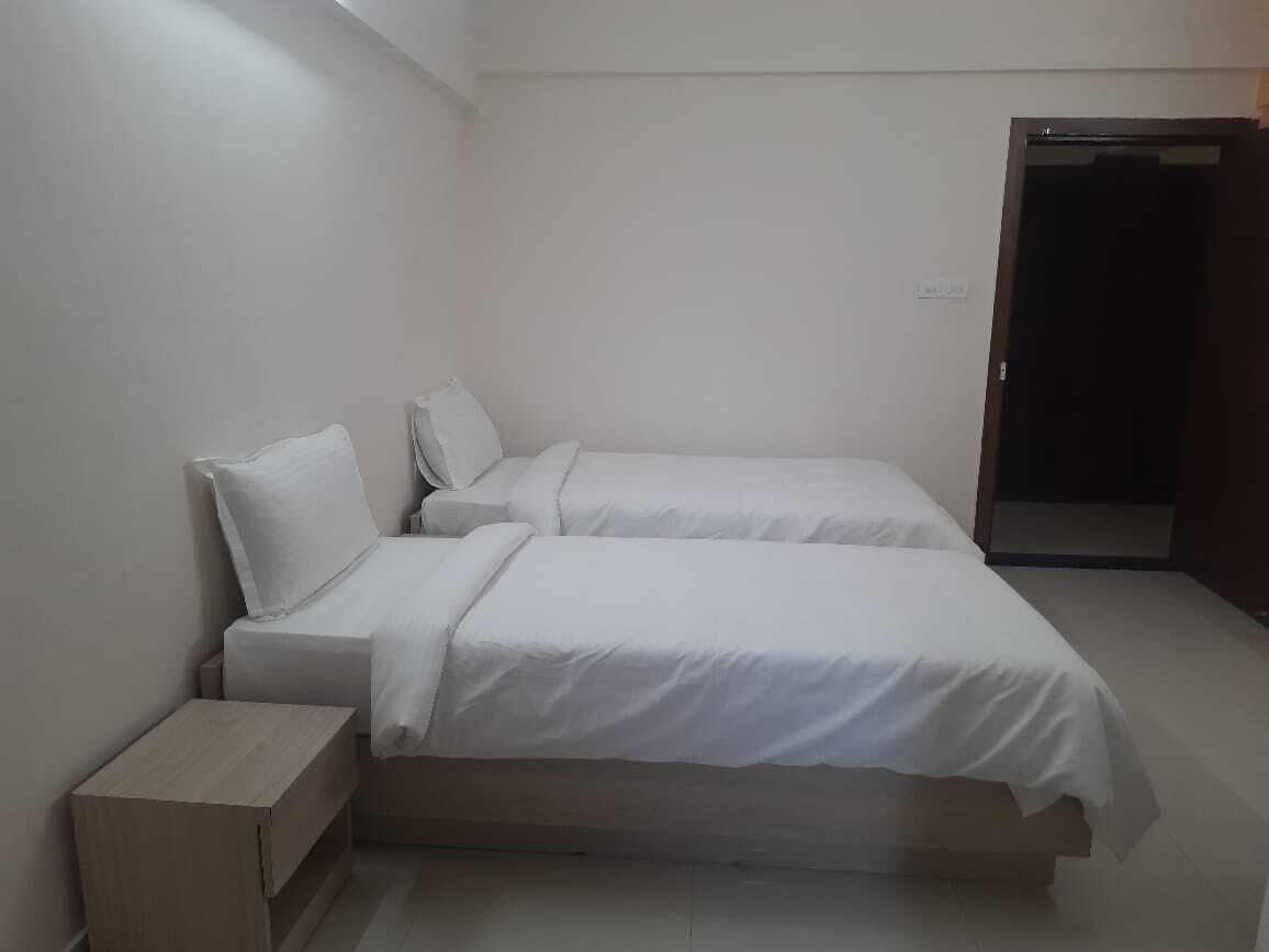Una habitación pequeña y limpia con dos camas individuales, una mesita de noche de madera y una puerta que da a otra habitación.